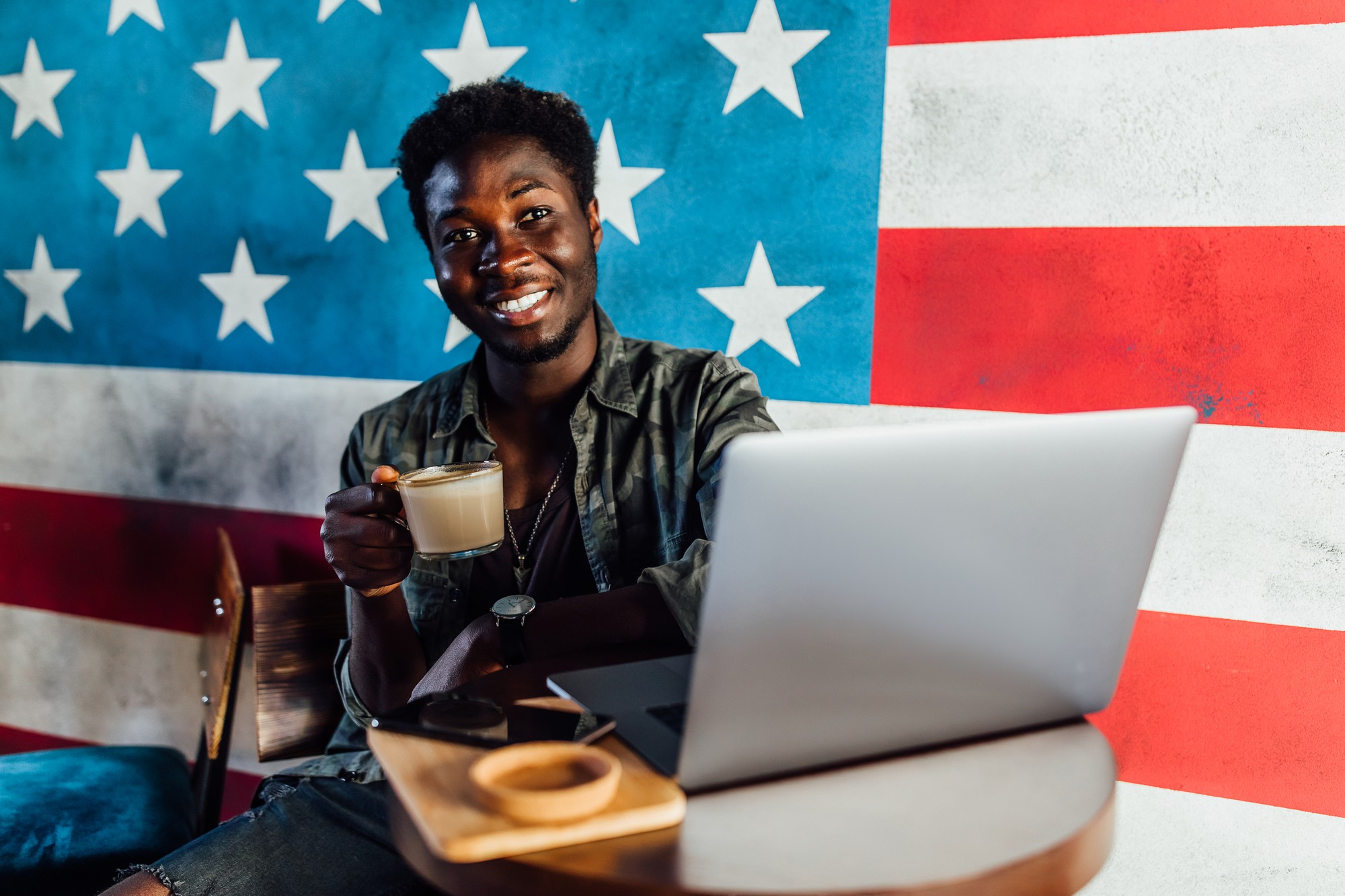 A man sitting drinking coffee with the USA flag hanging on a wall behind him | High Paying Jobs in the USA for Immigrants | School Infospot